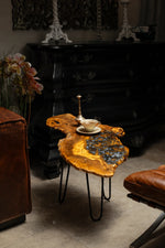 AI-styled view of Zitun Atelier table 037, an olive wood resin side table with blue pebble resin, in a dark interior beside leather seating.