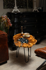AI-styled view of Zitun Atelier table 044, an olive wood resin side table with black resin nautical charms, in a dark interior beside leather seating.