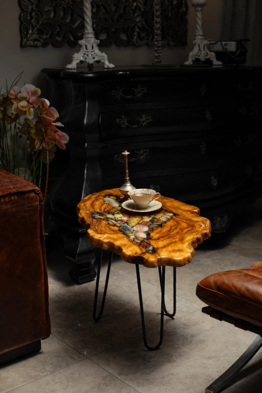 AI-styled view of Zitun Atelier table 047, an olive wood resin side table with sea shell pebble resin, in a dark interior beside leather seating.