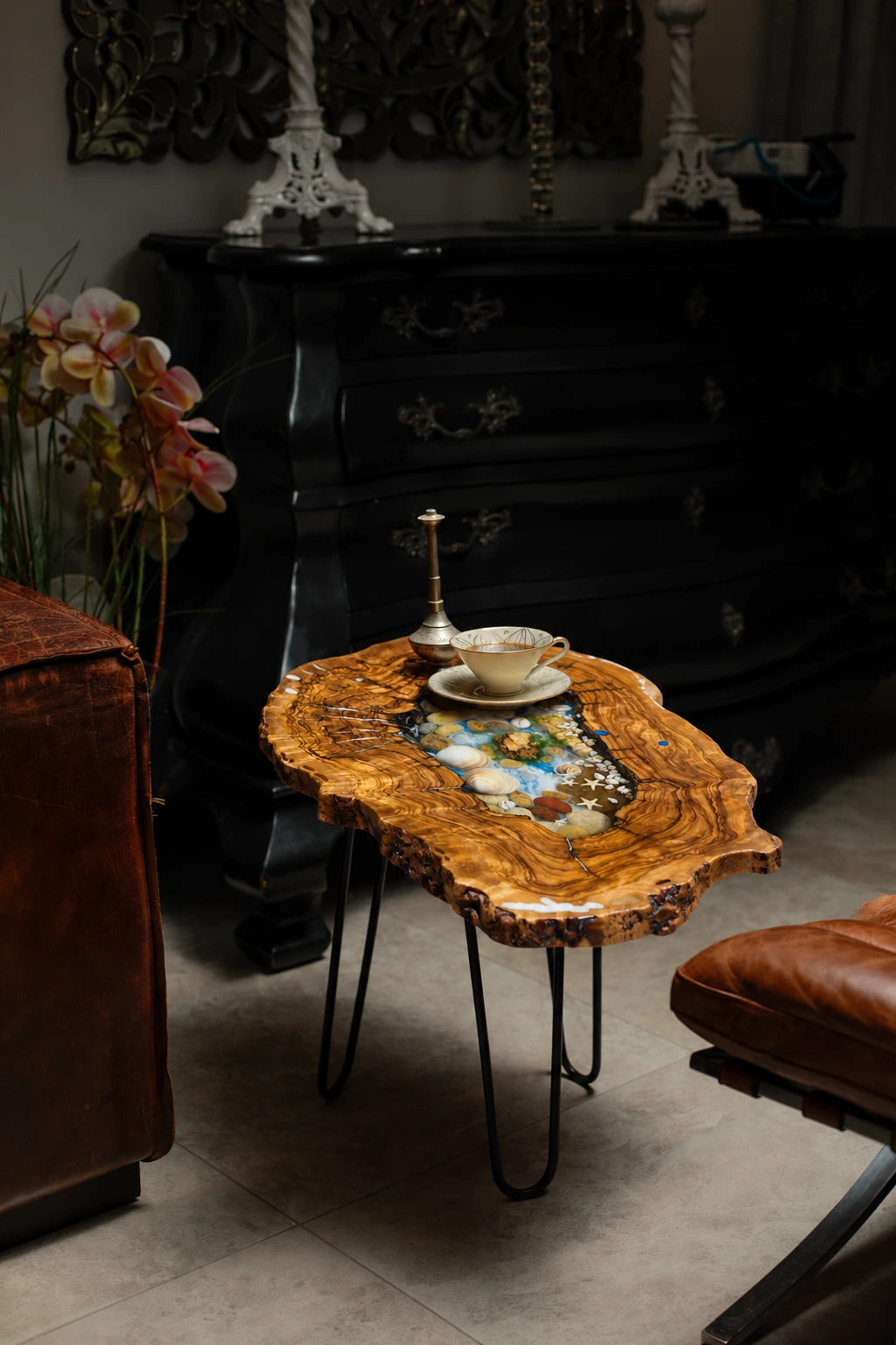 AI-styled view of Zitun Atelier table 052, an olive wood resin side table with sea shell pebble resin, in a dark interior beside leather seating.