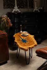 AI-styled view of Zitun Atelier table 079, an olive wood resin side table with blue starburst resin, in a dark interior beside leather seating.